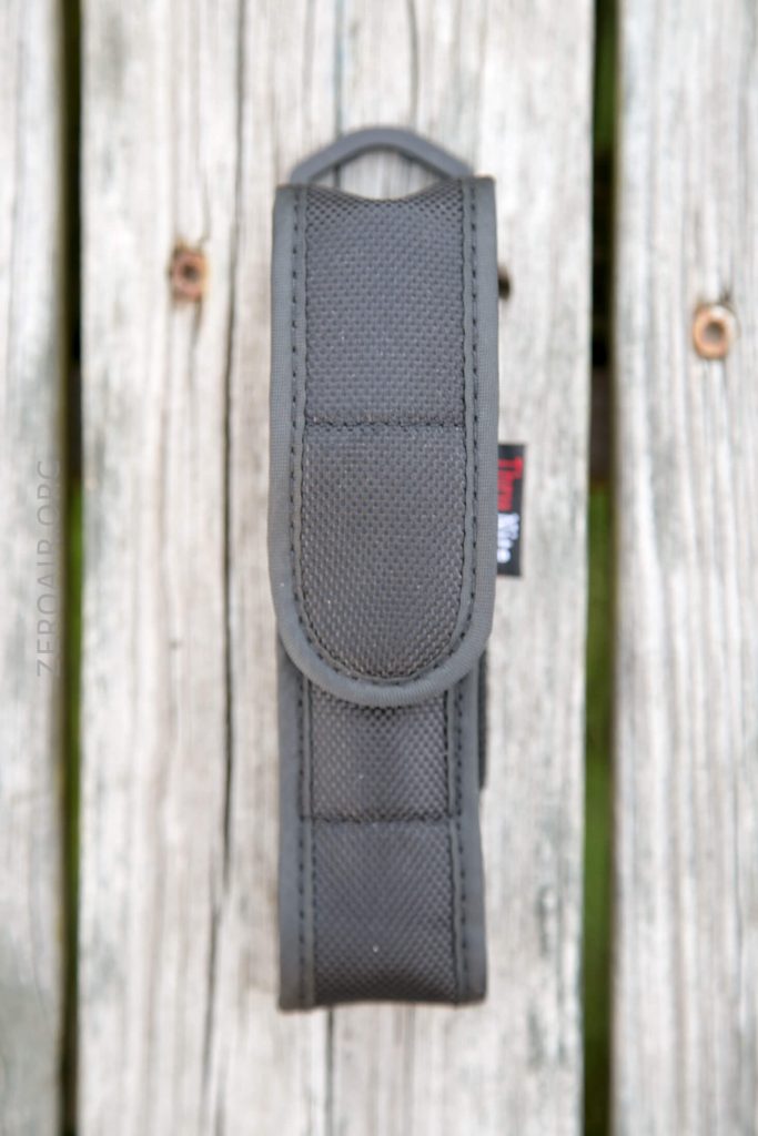 A black nylon pouch with a Velcro flap is shown vertically on a weathered wooden surface. The pouch has reinforced stitching and a small black and red tag on the side.