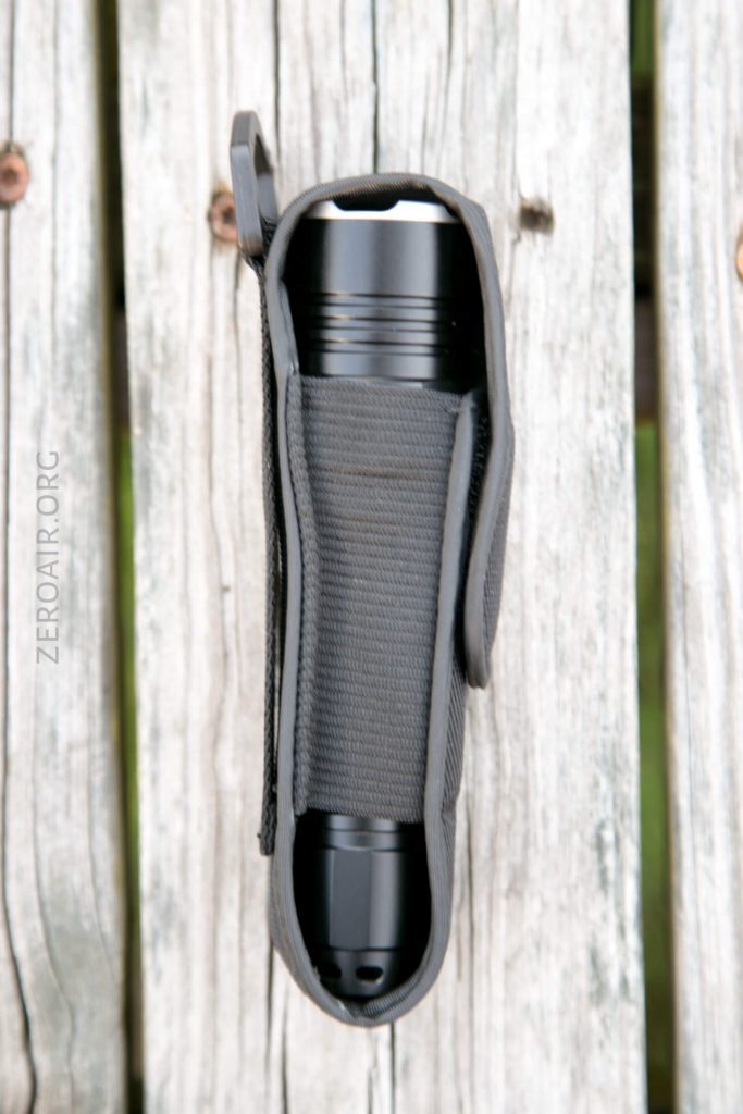 A black flashlight in a gray fabric holster, placed vertically on a weathered wooden surface. The holster is partially open, revealing the flashlight inside. The background consists of faded wooden planks.