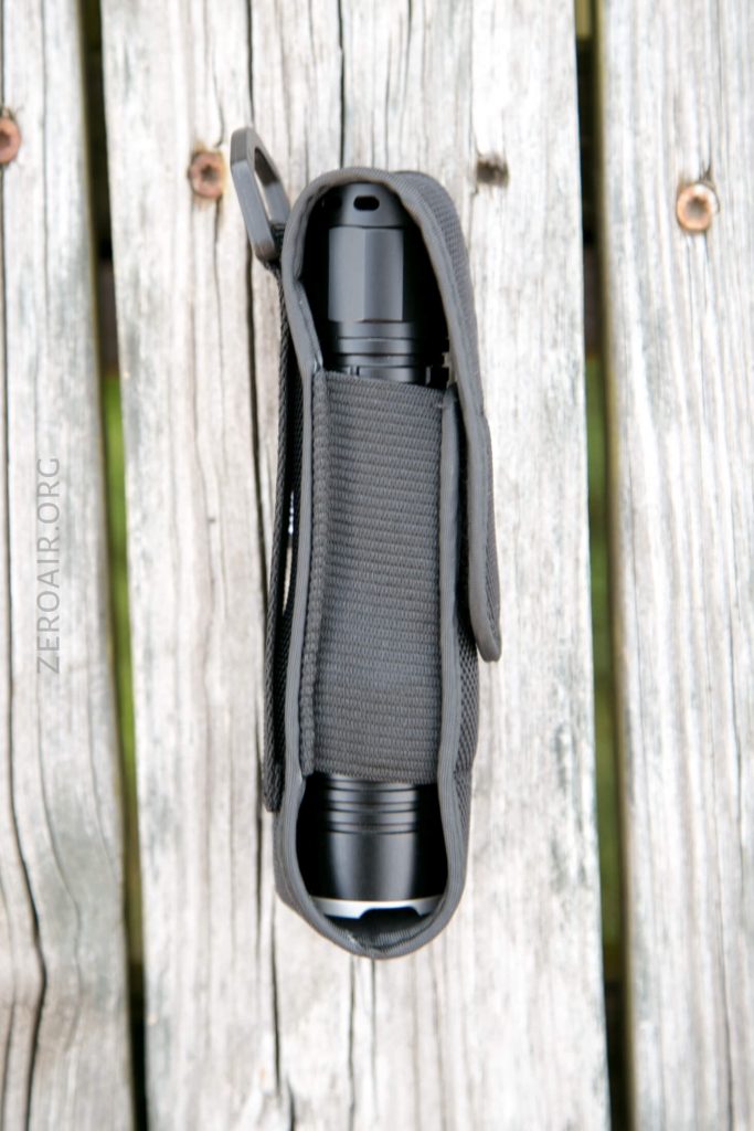 A black flashlight secured in a black fabric holster with a Velcro flap, resting on weathered wooden planks.