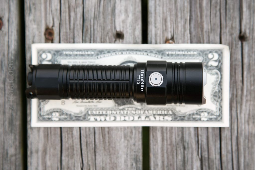 A black ThruNite TN10 flashlight is placed on top of a two-dollar bill, both resting on a weathered wooden surface.