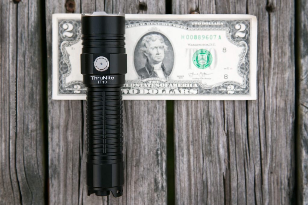 A black ThruNite TT10 flashlight is placed on top of a two-dollar bill, both lying on a weathered wooden surface. The flashlight is positioned vertically across the bill.