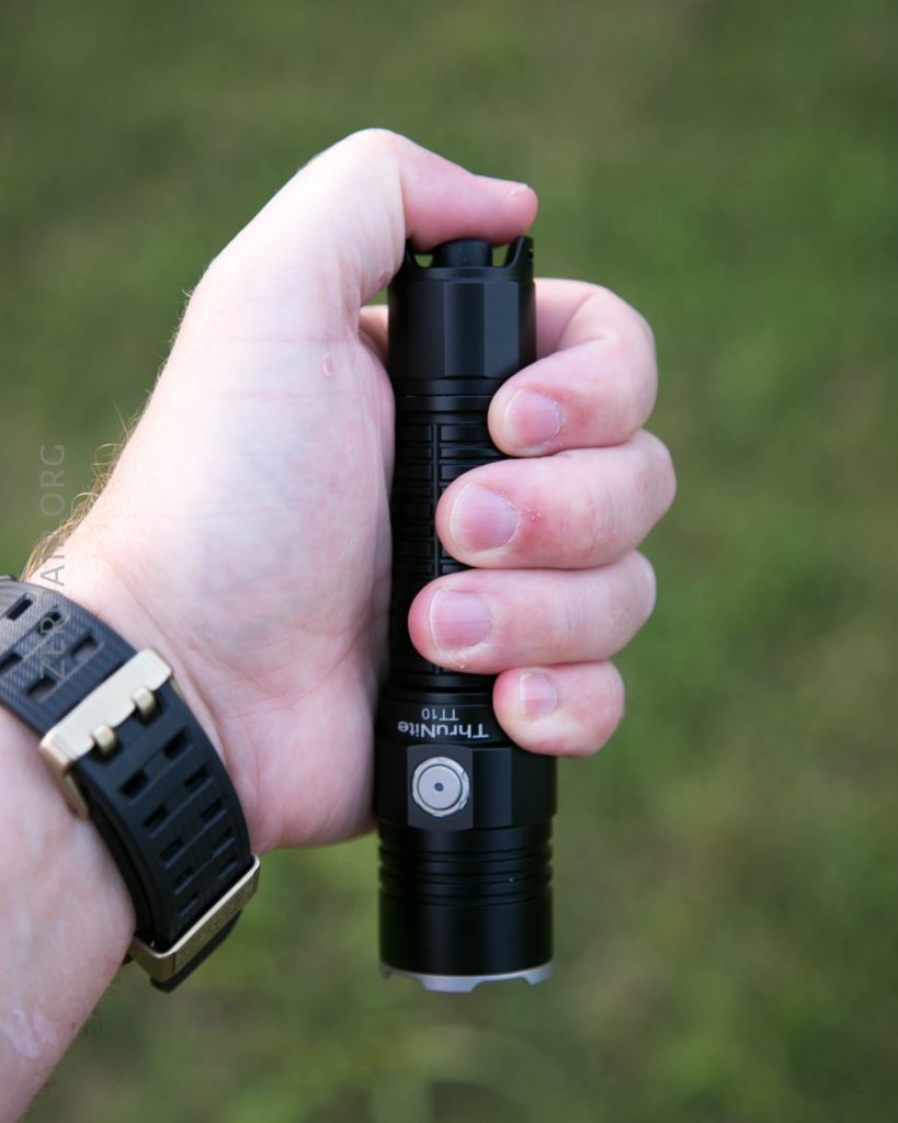 A person wearing a black wristwatch holds a black ThruNite T11T flashlight in their right hand against a blurred green background.