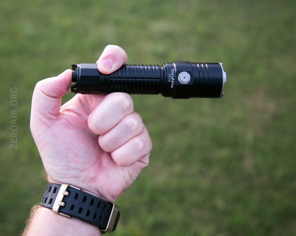 A person holding a black flashlight horizontally in their right hand against a blurred grassy background. The person is wearing a black wristwatch with a gold buckle.
