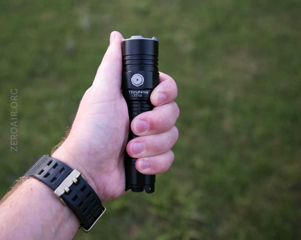 A person holding a black ThruNite TT10 flashlight in their right hand outdoors, with a grassy background visible and wearing a black wristwatch.