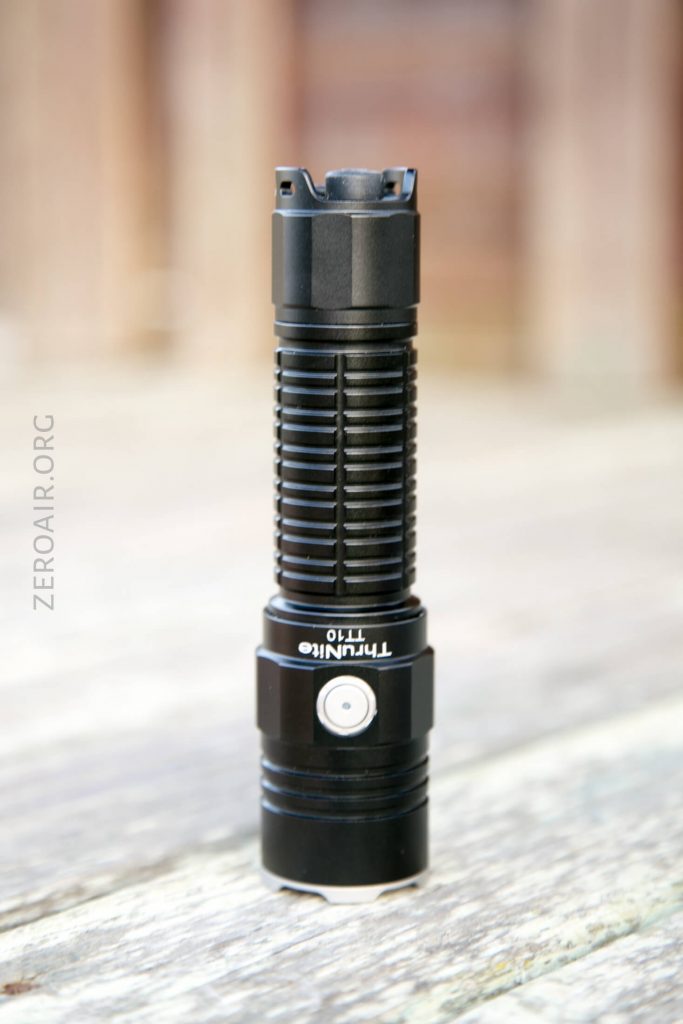 A black Thrunite T1T0 flashlight stands upright on a wooden surface with a blurred background. The flashlight has a ribbed grip and a prominent silver power button.