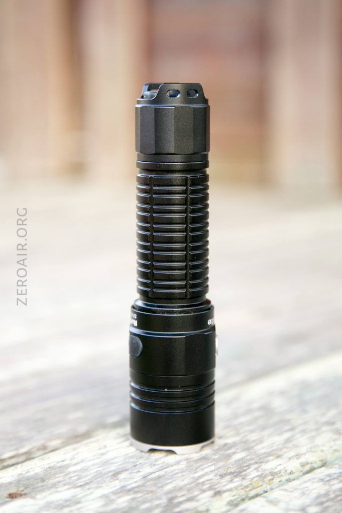 A black, cylindrical flashlight with a ribbed grip stands upright on a wooden surface. The background is blurred, and ZEROAIR.ORG is faintly visible along the left side.