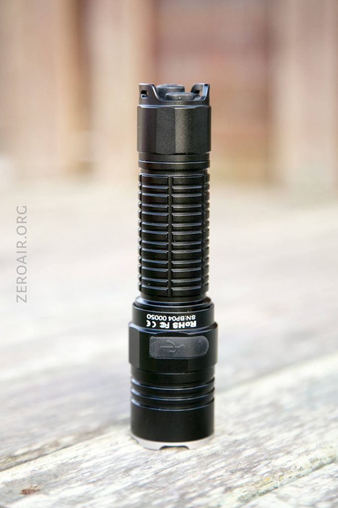 A black, cylindrical flashlight with ridged grip details stands upright on a light-colored wooden surface. The background is softly blurred, with vertical brown panels visible.