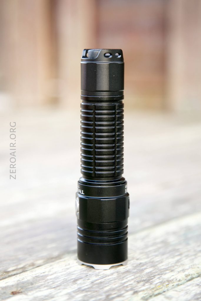 A black, cylindrical flashlight stands upright on a wooden surface. The flashlight has a ribbed grip and a flat base, with a blurred background. The text ZEROAIR.ORG is vertically visible to the left.