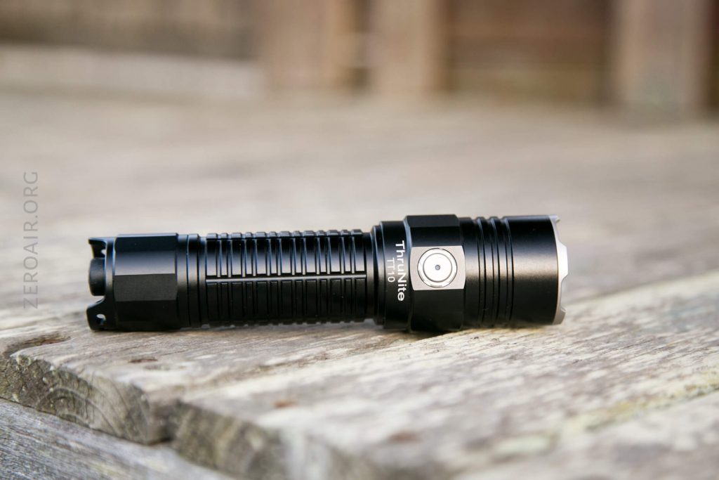 A black, cylindrical flashlight with ribbed grip and side button lies on a weathered wooden surface. The flashlight is positioned horizontally with the lens facing right.