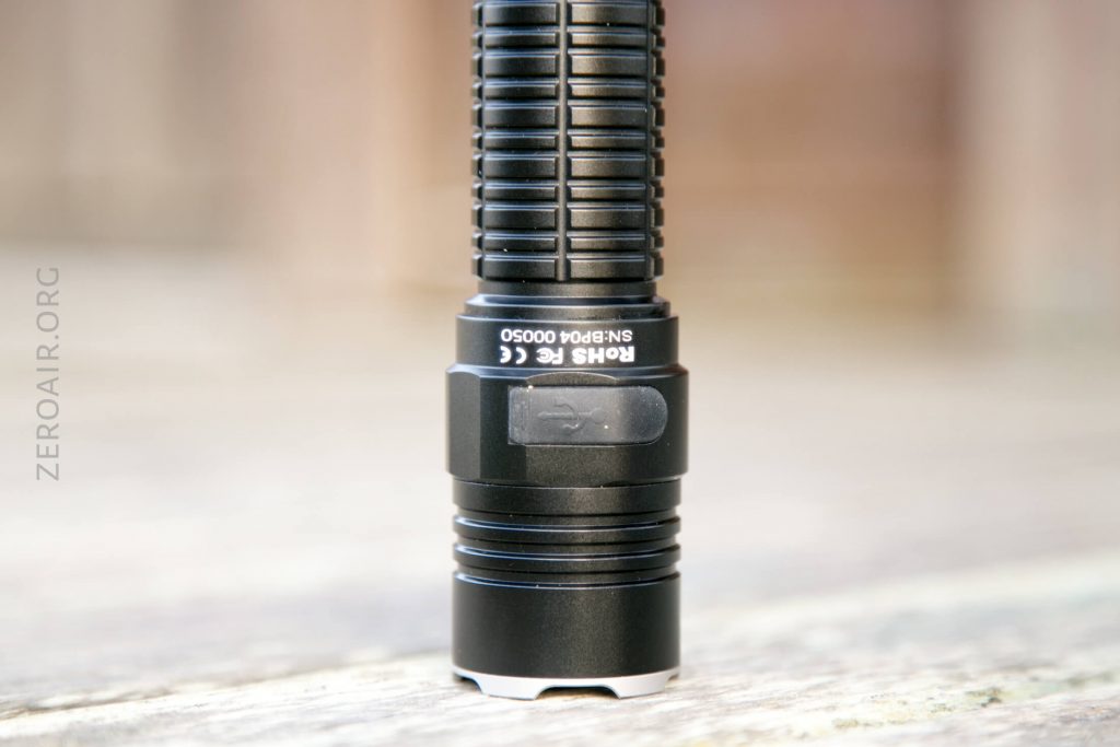 A close-up of a black, cylindrical flashlight standing upright on a blurred surface. The flashlight has ridged grip details and visible text that includes “ROHS” and “FC.” The background is out of focus.