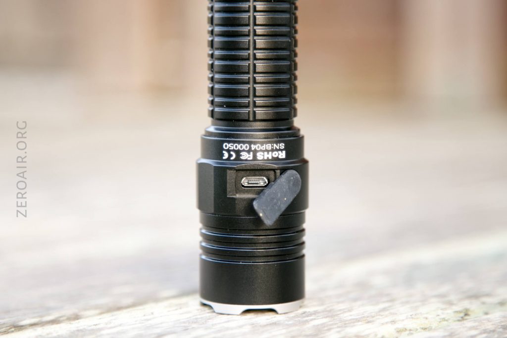 A close-up of a black flashlight standing upright on a blurred surface, showing a rubber flap open to reveal a micro USB charging port on the body of the flashlight.