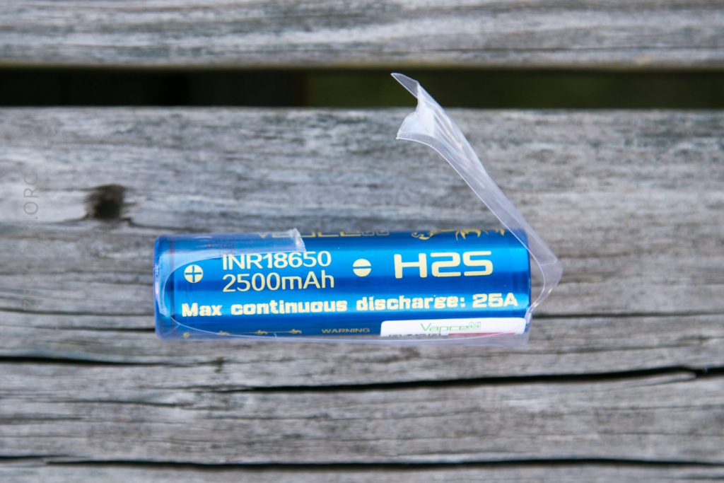A blue cylindrical lithium-ion battery labeled INR18650, 2500mAh, and H2S, with clear plastic partially peeling off, resting on a weathered wooden surface.
