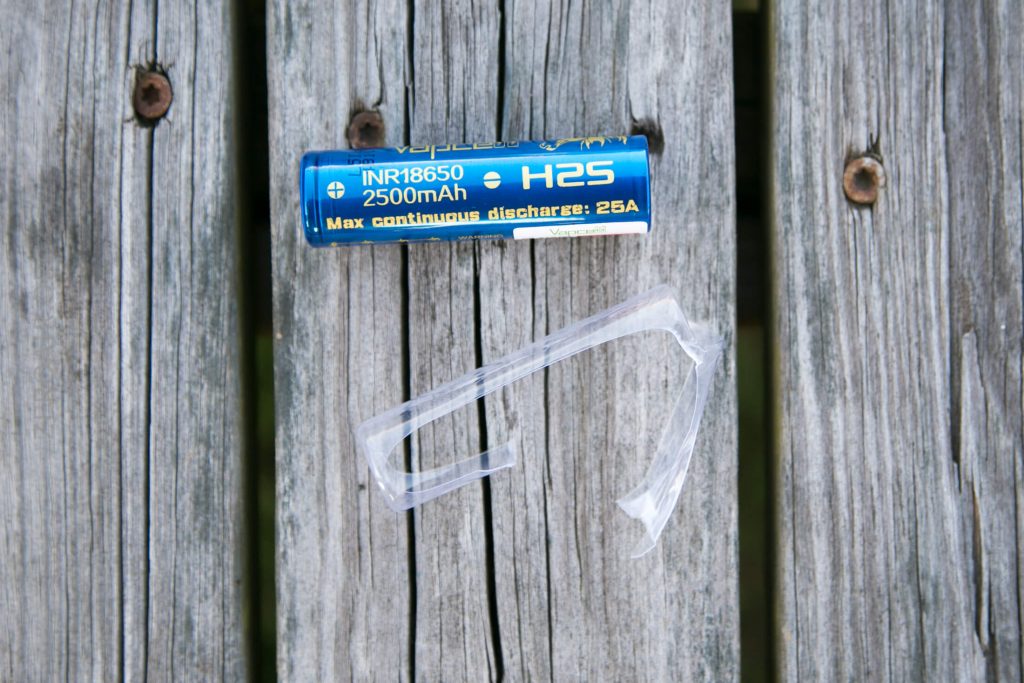 A blue cylindrical 2500mAh H2S INR18650 battery and a broken clear plastic battery holder are placed on a weathered wooden surface with visible gaps between the planks.