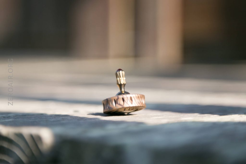 A metal spinning top with engraved patterns is spinning on its side outdoors on a wooden surface, with sunlight casting shadows and a blurred background.