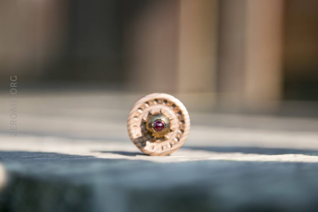 A close-up photo of a cylindrical wooden object placed upright on a stone surface, with a small red gemstone set in the center facing the camera. The background is blurred.