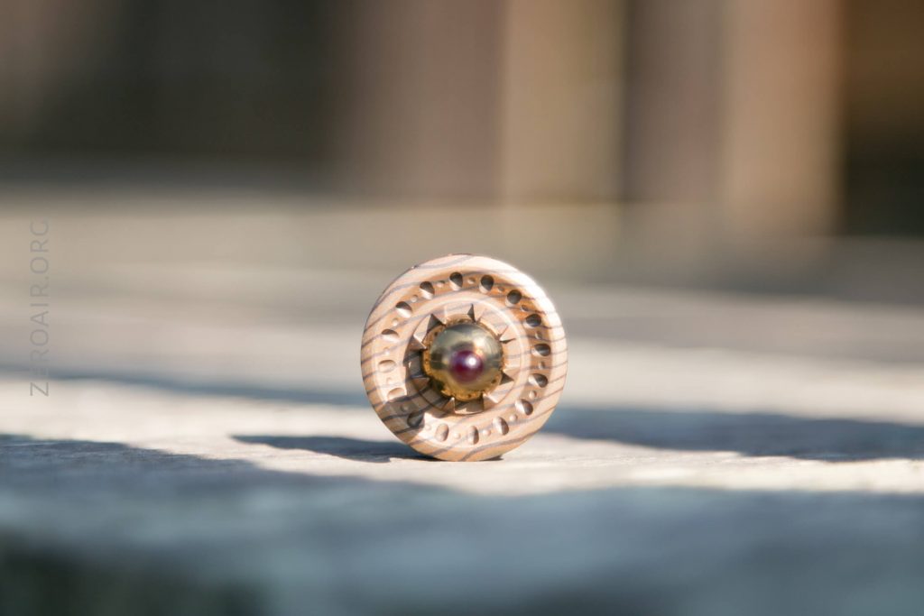 A round, carved wooden object with concentric ring patterns and a small metallic or beaded centerpiece rests on a sunlit, blurred surface. The background features vertical, out-of-focus lines.