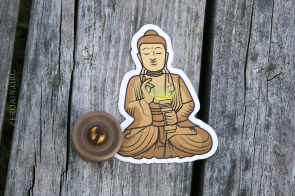 A sticker depicting a seated Buddha figure is placed on weathered wooden boards. A spinning top is in motion near the bottom left of the sticker. The word ZEROAIR.ORG is visible on the wood.