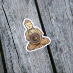 A sticker of a seated Buddha figure is placed on weathered wooden planks, with a brown decorative round object attached to the center of the figure.