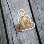 A sticker depicting a meditating Buddha figure sits on a weathered wooden surface next to a round, metallic pin backing.