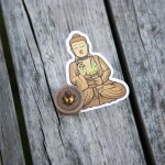 A sticker of a meditating Buddha is placed on a weathered wooden surface. In front of the sticker, there is a small transparent capsule containing a golden object.