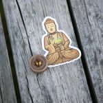 A sticker of a meditating Buddha and a small round object with a metallic center sit on weathered wooden planks.