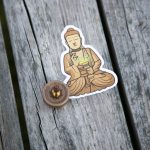 A sticker of a meditating Buddha holding a green bowl is placed on weathered wooden boards. Nearby, a small brown spinning top with a gold tip rests on the wood.