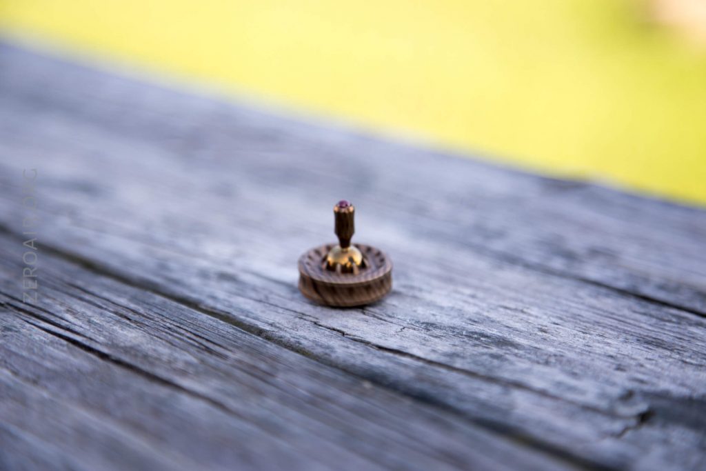 A small spinning top made of wood and metal is placed upright on a weathered wooden surface outdoors, with a blurred green background.