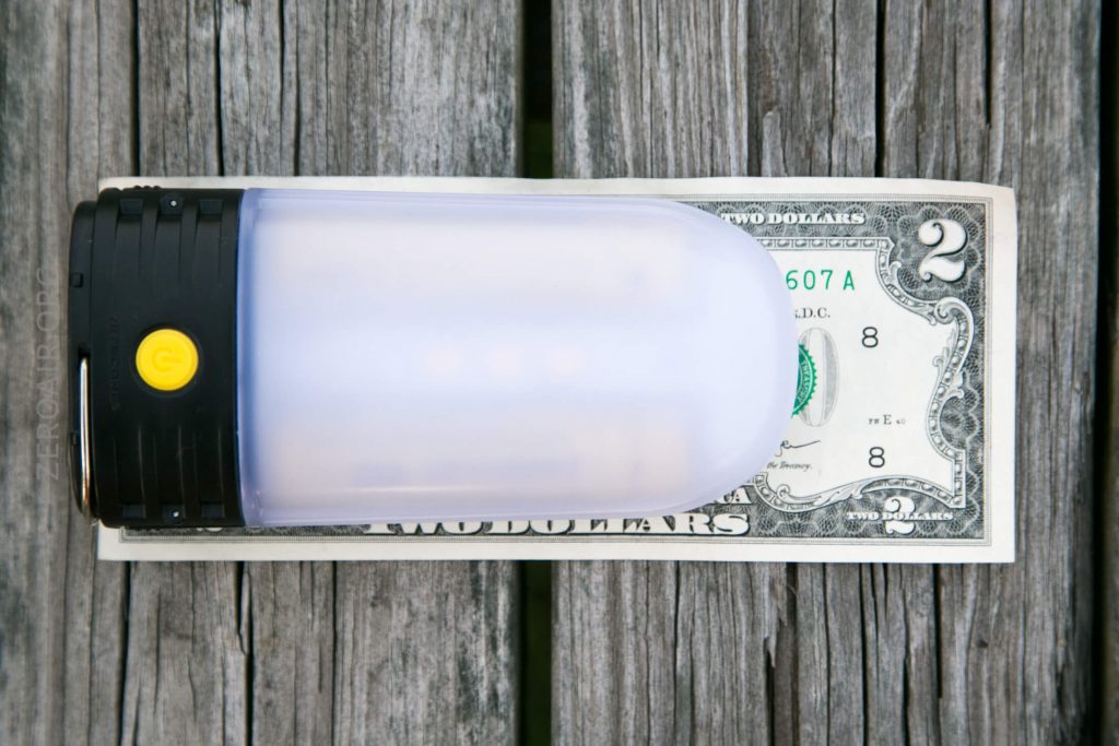 A rectangular LED camping lantern with a yellow power button is placed on top of a two-dollar bill on a weathered wooden surface.