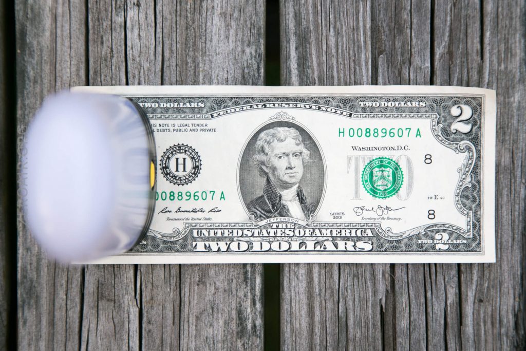 A U.S. two-dollar bill lies flat on a wooden surface, partially covered on the left side by a cylindrical, translucent white object, creating a blurred effect over that area.