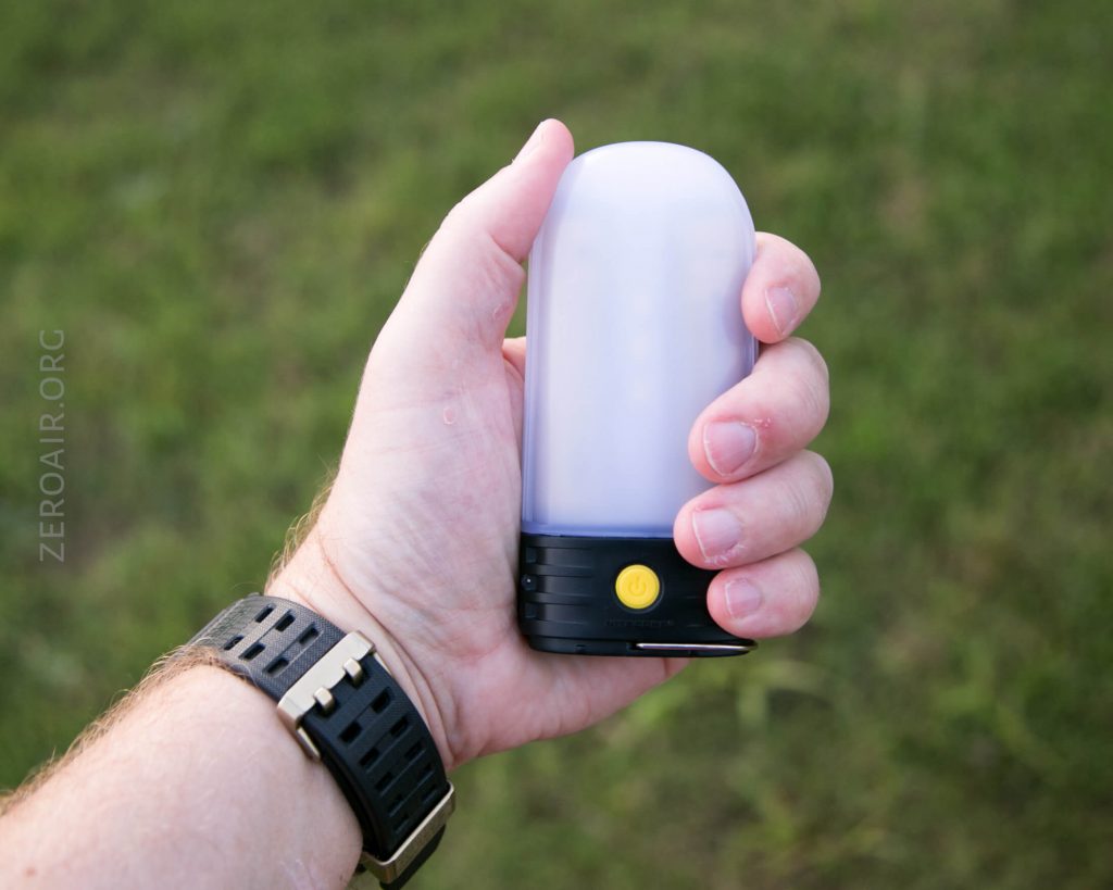 A person’s hand holding a small, portable lantern with a white frosted dome and a yellow power button. The person is wearing a black watch. The background is grassy.