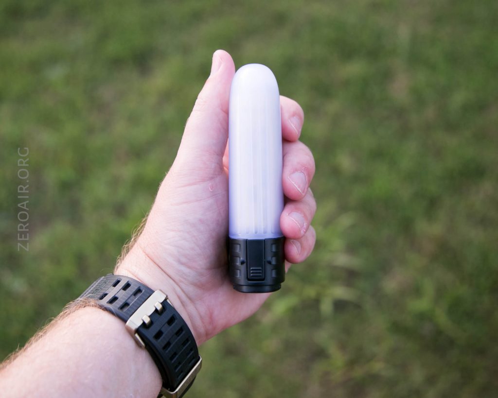 A person with a black wristwatch holds a small, cylindrical, white and black object outdoors over grass. The object appears to be a portable light or flashlight.
