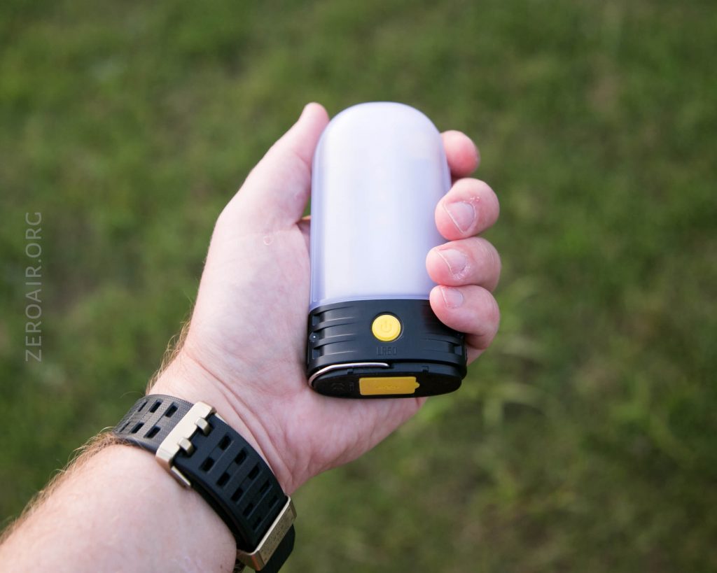 A person holds a small, cylindrical LED lantern with a yellow power button, black base, and frosted white top. The person wears a black watch with a gold buckle. The background is grassy and outdoors.