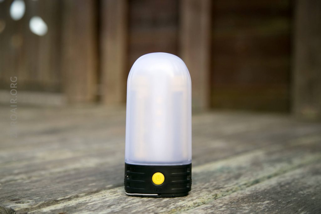 A small, portable LED lantern with a white frosted dome and a black base featuring a single yellow button sits on a weathered wooden surface.