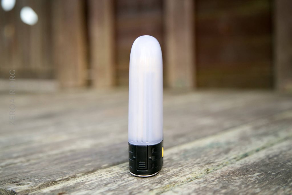 A small, cylindrical LED lamp with a white diffusing top and a black base is standing upright on a wooden surface, with a blurred wooden background.