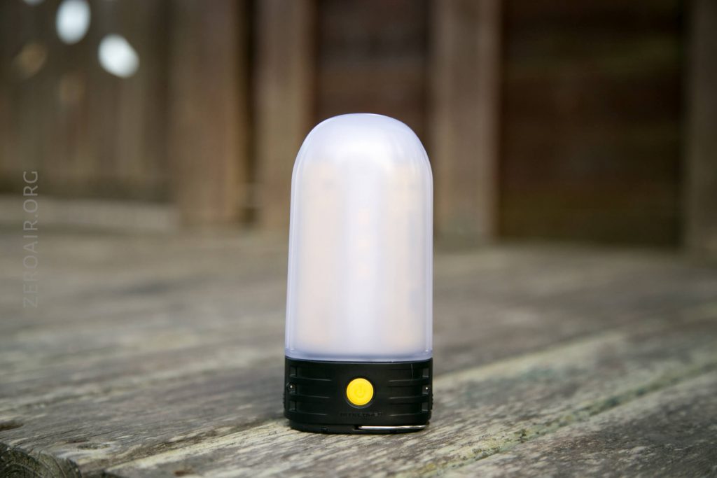 A portable LED lantern with a frosted dome-shaped cover and a black base featuring a yellow power button sits on a weathered wooden surface.