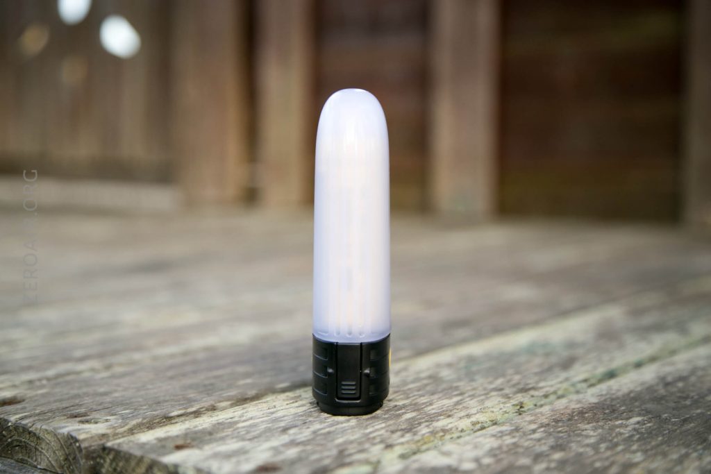 A small, cylindrical, battery-powered LED lantern with a white top and black base is standing upright on a weathered wooden surface outdoors.
