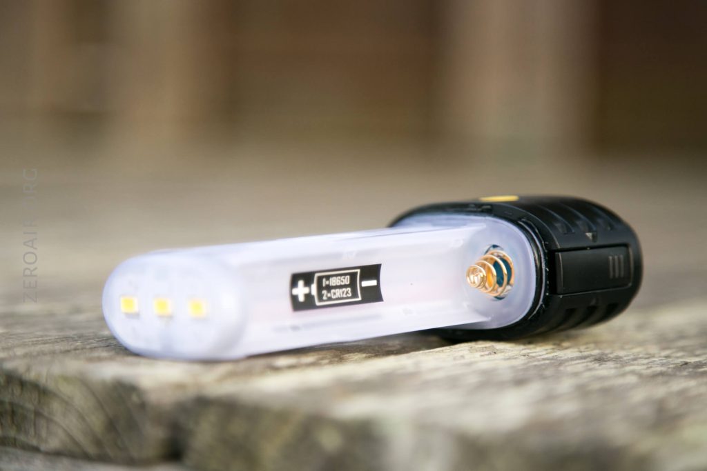 A close-up of a portable flashlight lying on a wooden surface, showing its clear body, three small LED lights, a label with symbols, and a metallic contact point.