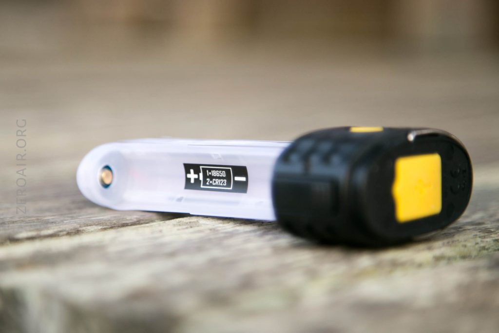 A close-up of a small flashlight lying on a wooden surface. The flashlight has a transparent body, a yellow button on the black cap, and a visible battery orientation label.