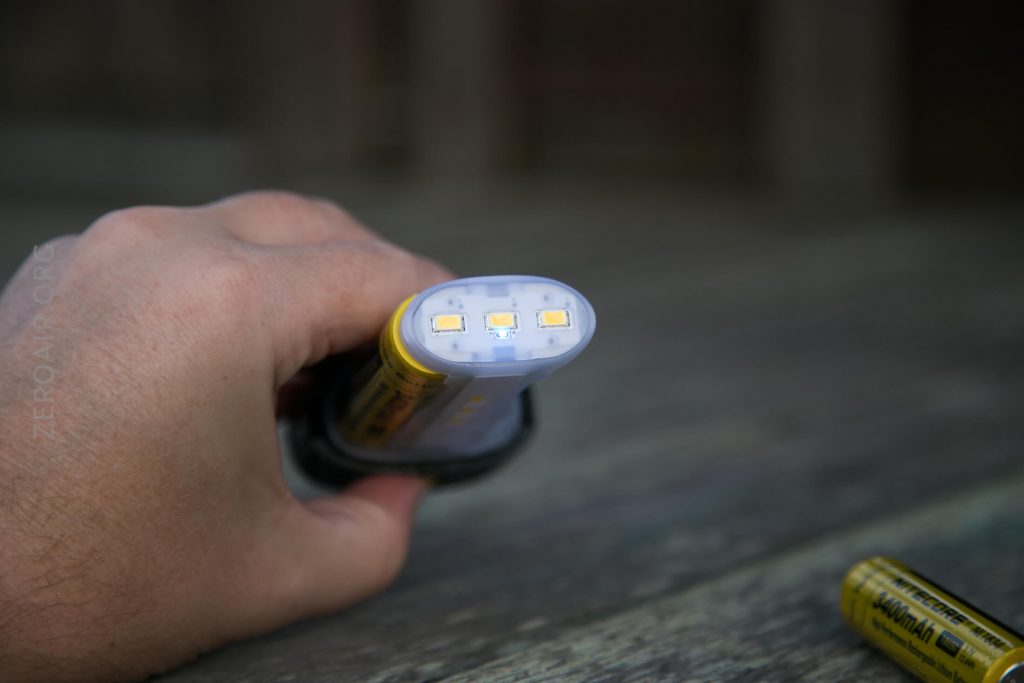 A hand holds a cylindrical battery with three small yellow LED lights on one end. Another battery lies on a wooden surface in the background.