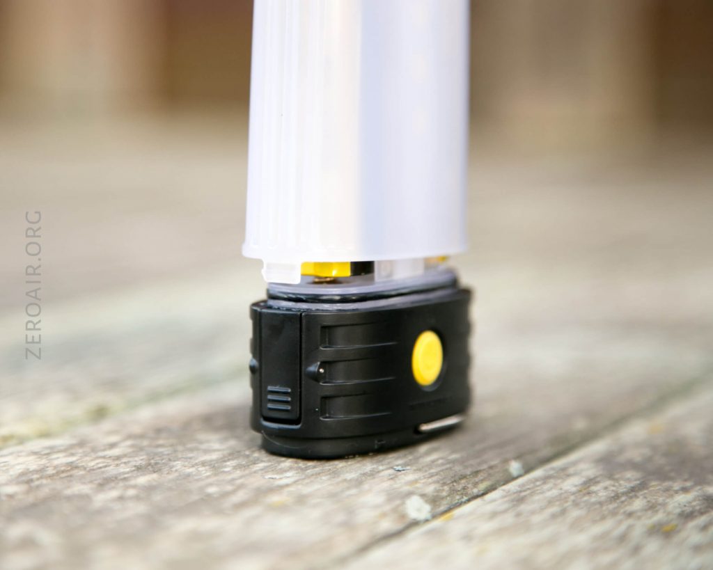 A close-up of the base of a portable LED lantern with a black and yellow control unit, standing on a wooden surface. The translucent body of the lantern is partially visible.