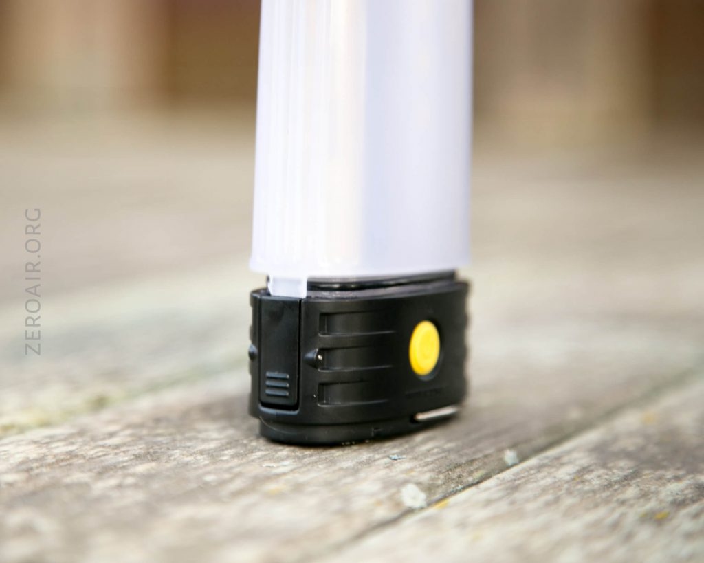 A close-up of the base of a portable LED lantern with a black and yellow button, resting on a weathered wooden surface. The lantern has a cylindrical white body.