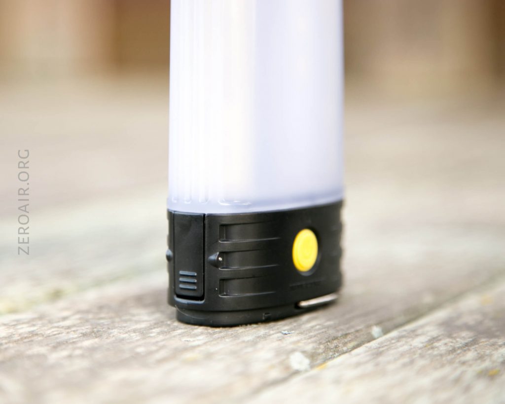 A close-up of the base of a cylindrical light or lantern with a black bottom, a yellow button, a small port, and a semi-transparent white top, placed on a weathered wooden surface.