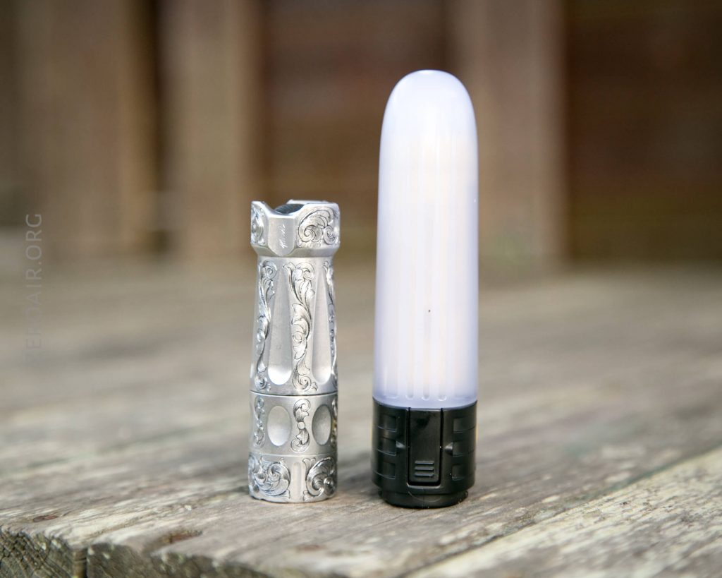 A small, ornate metal flashlight stands next to a taller, pill-shaped LED light with a black base on a weathered wooden surface.