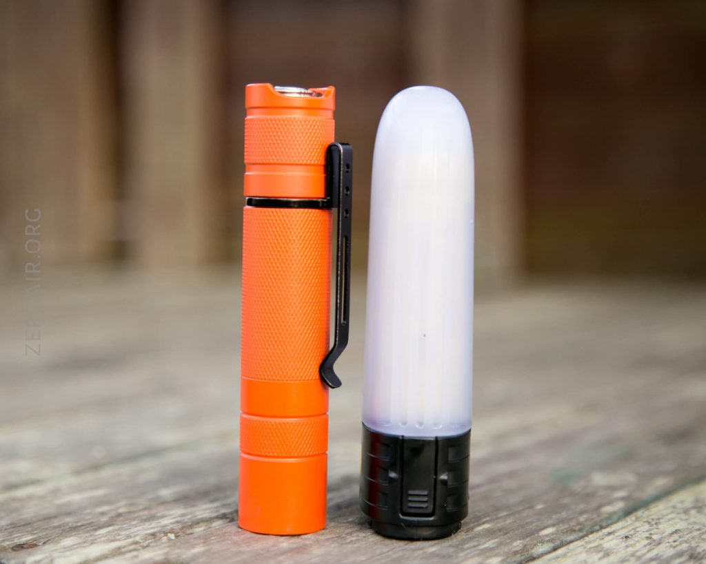 An orange flashlight with a black clip stands upright next to a white, rounded diffuser attachment on a wooden surface with a blurred background.