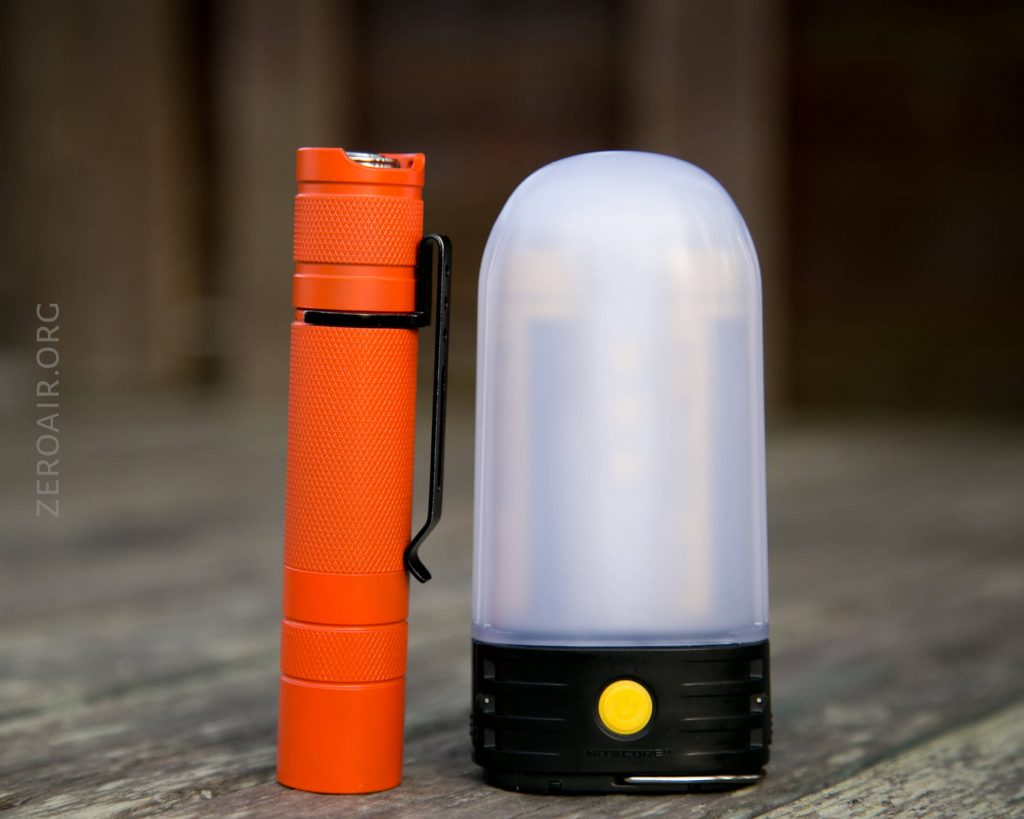 An orange flashlight stands upright next to a small, dome-shaped LED lantern with a frosted white top and a black base featuring a yellow power button. Both objects are on a wooden surface.