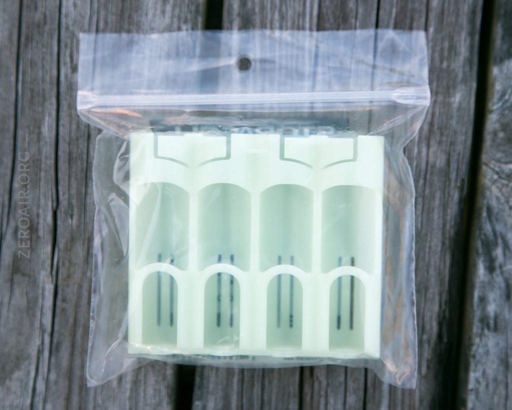 A translucent plastic battery holder is placed inside a clear resealable plastic bag, resting on a weathered wooden surface. The holder has four slots for cylindrical batteries.