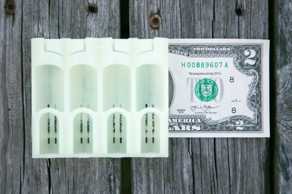 A white four-slot AA battery holder is placed next to a U.S. two-dollar bill on a weathered wooden surface. The items are arranged side by side for size comparison.