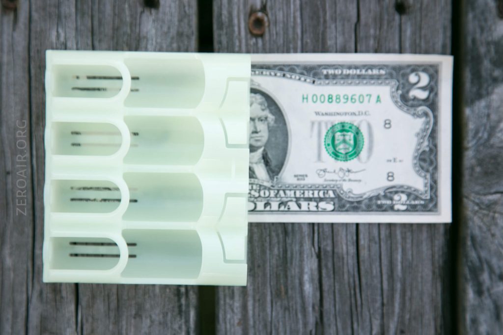 A white plastic object with several rectangular slots is placed on top of a US two-dollar bill, both resting on weathered wooden boards.