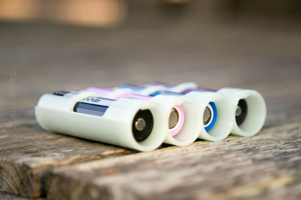 Four cylindrical batteries in white plastic holders are arranged in a row on a wooden surface. The batteries have visible colored rings—black, pink, blue, and purple—near the terminals.