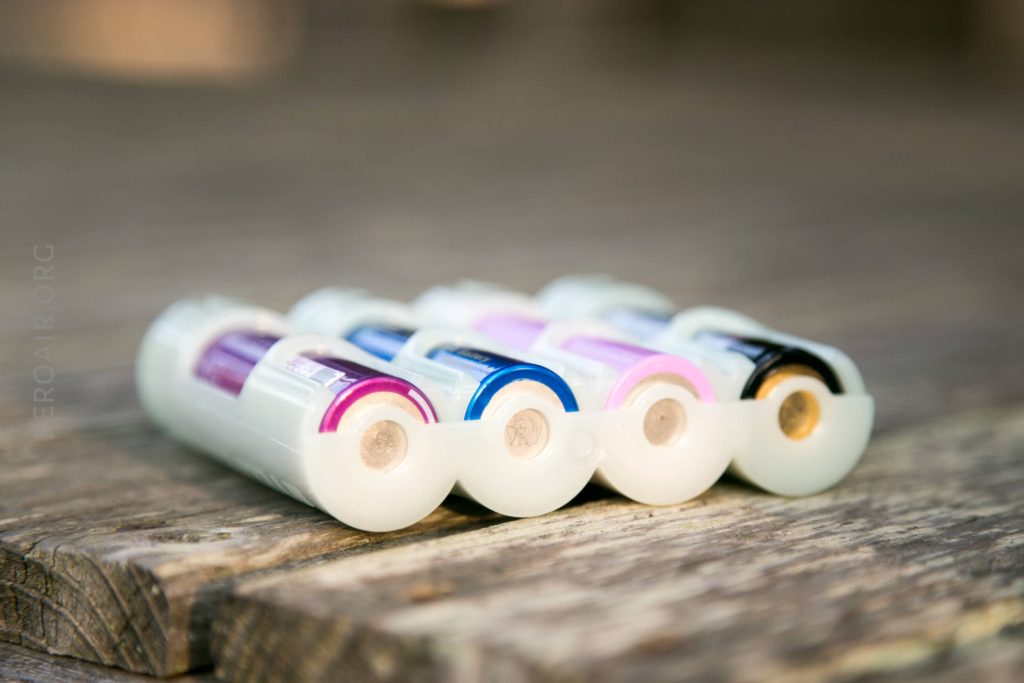 Four AA batteries with colored labels are arranged side by side in a white plastic holder, placed on a wooden surface. The photo is taken at close range, focusing on the battery terminals.